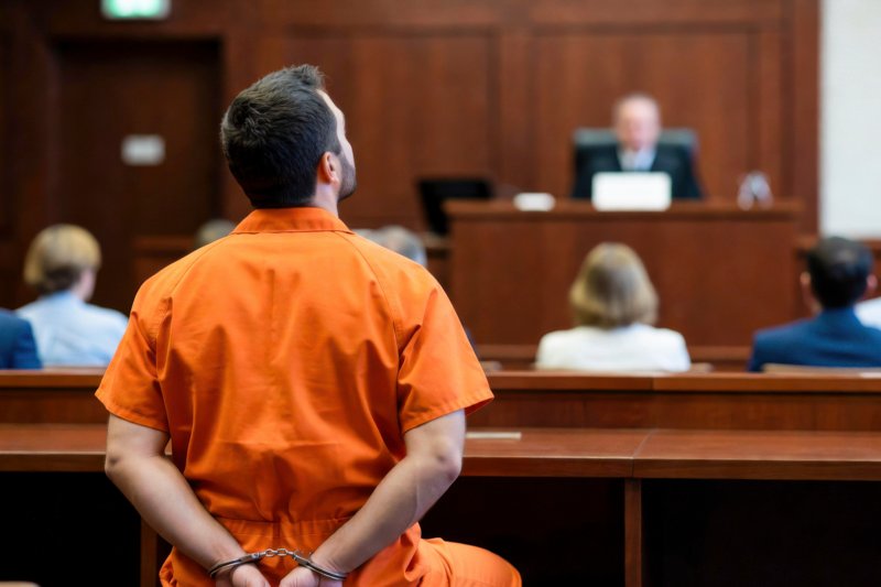A man in an orange jumpsuit and handcuffs sits with his back to